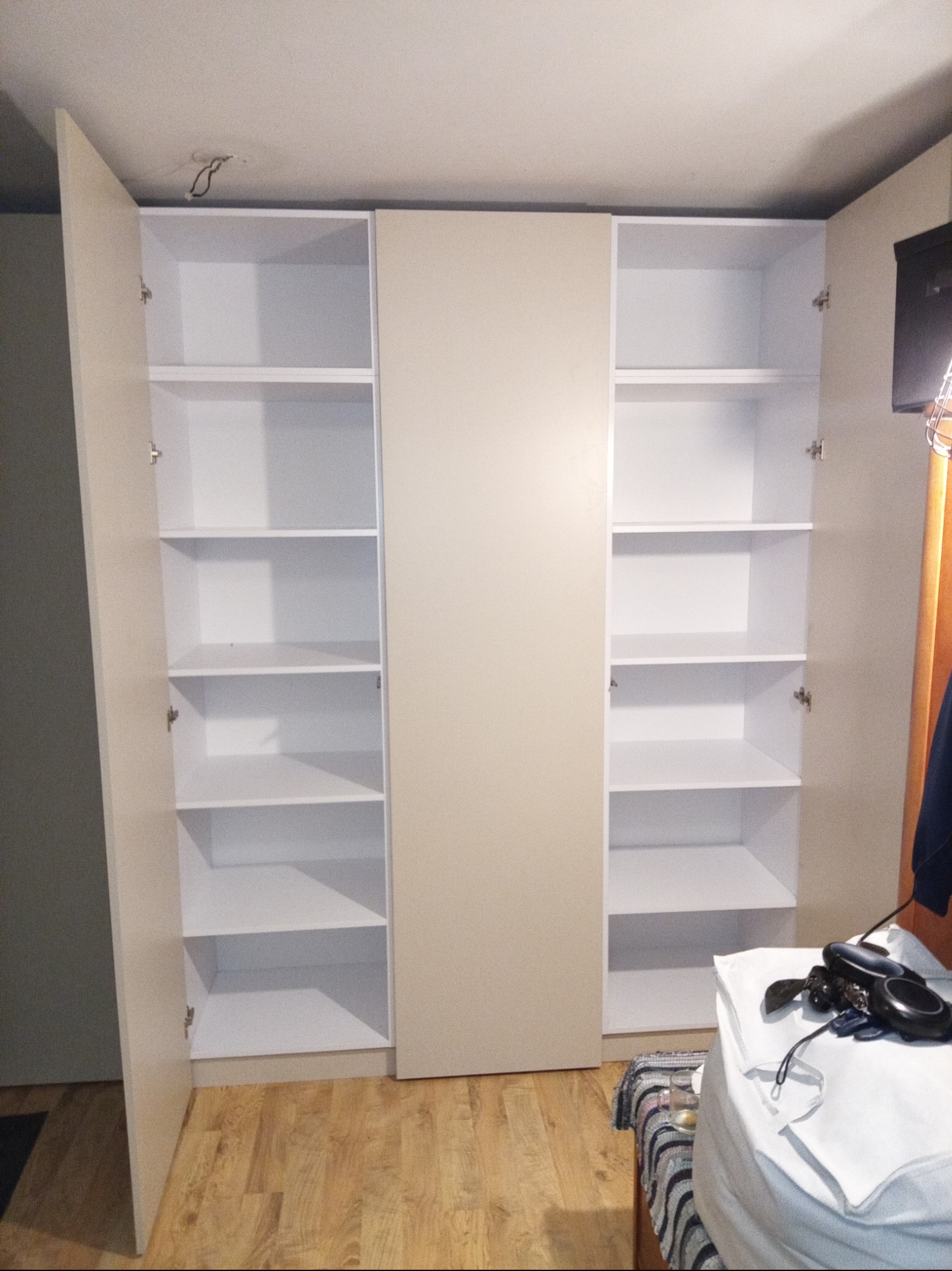 Trzydrzwiowa szafa z otwartymi drzwiami po bokach, ukazująca białe półki wewnątrz, z widoczną instalacją elektryczną na suficie i przedmiotami na łóżku w tle.