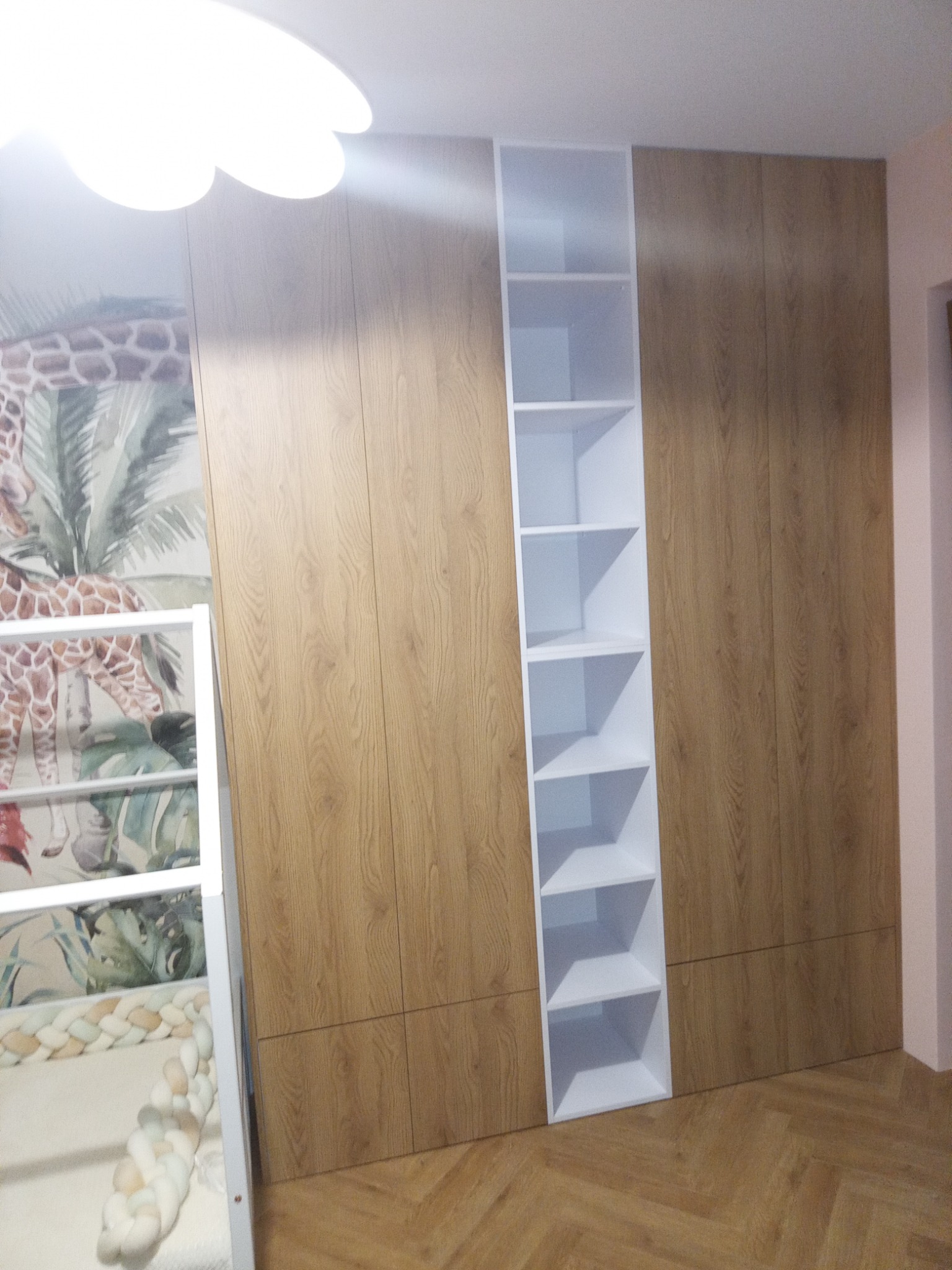 Dziecięca szafa wnękowa z drewnianymi frontami i białym regałem wbudowanym po lewej stronie, widoczny fragment łóżeczka dziecięcego z motywem zwierząt na tapecie w tle.