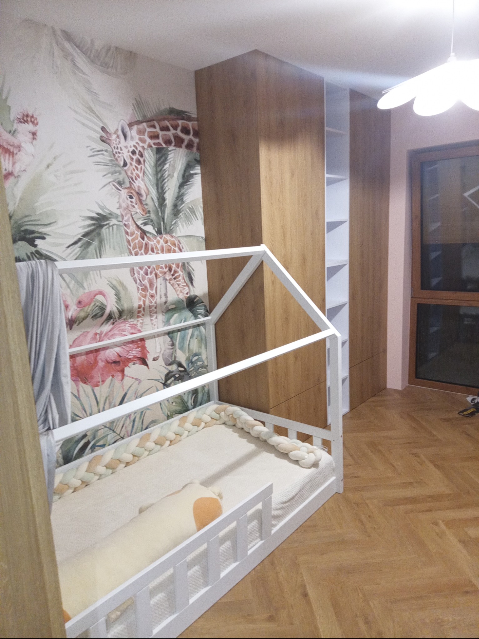 Pokój dziecięcy z białym łóżkiem w kształcie domku, plecionym ochraniaczem i poduszką, na tle szafy w kolorze drewna i tapety z motywem dżungli ze zwierzętami.