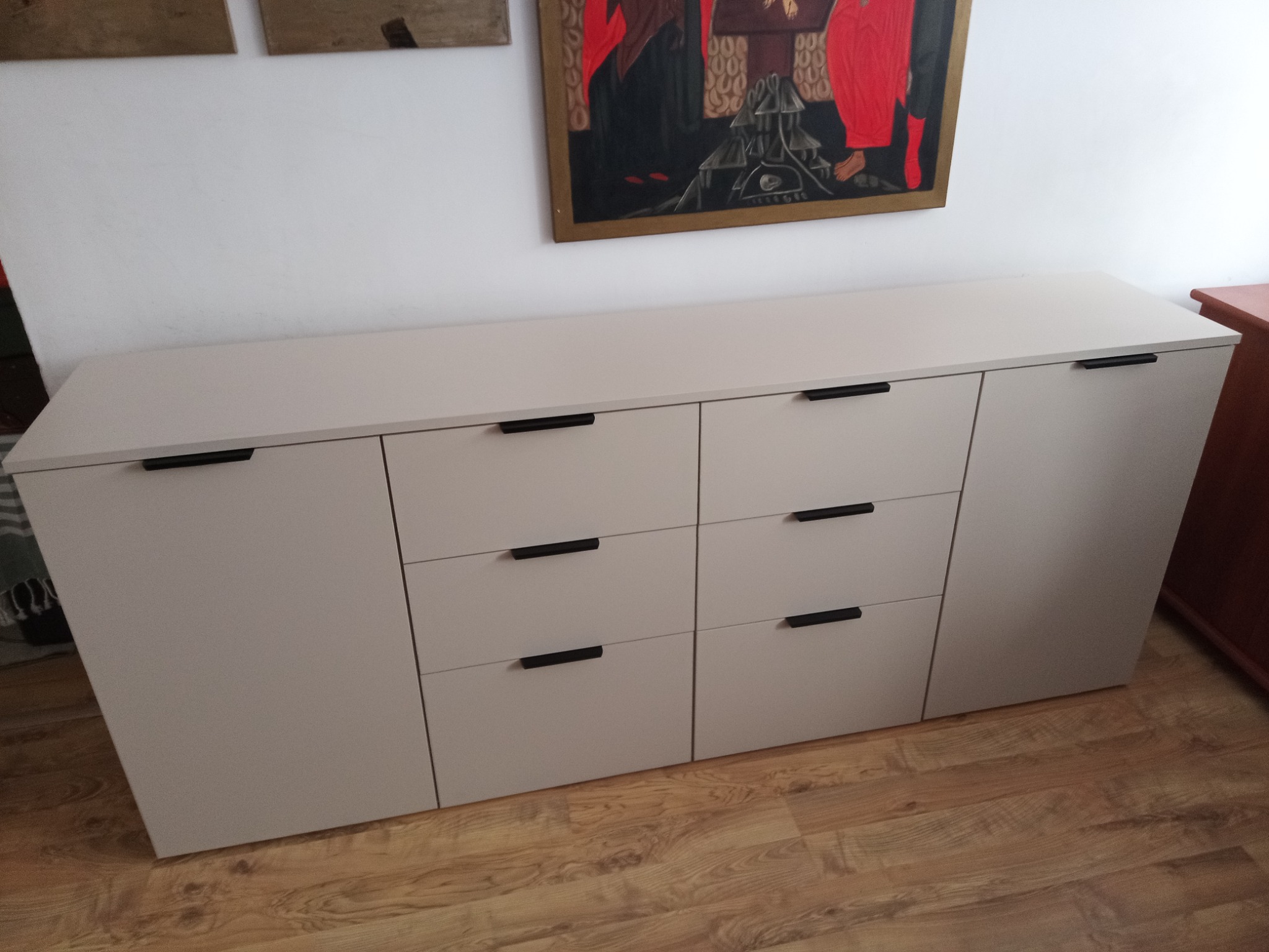 Jasnoszara komoda z czarnymi uchwytami na tle drewnianej podłogi i białej ściany z obrazem. Mebel z frontami bez zdobień, minimalistyczny design.