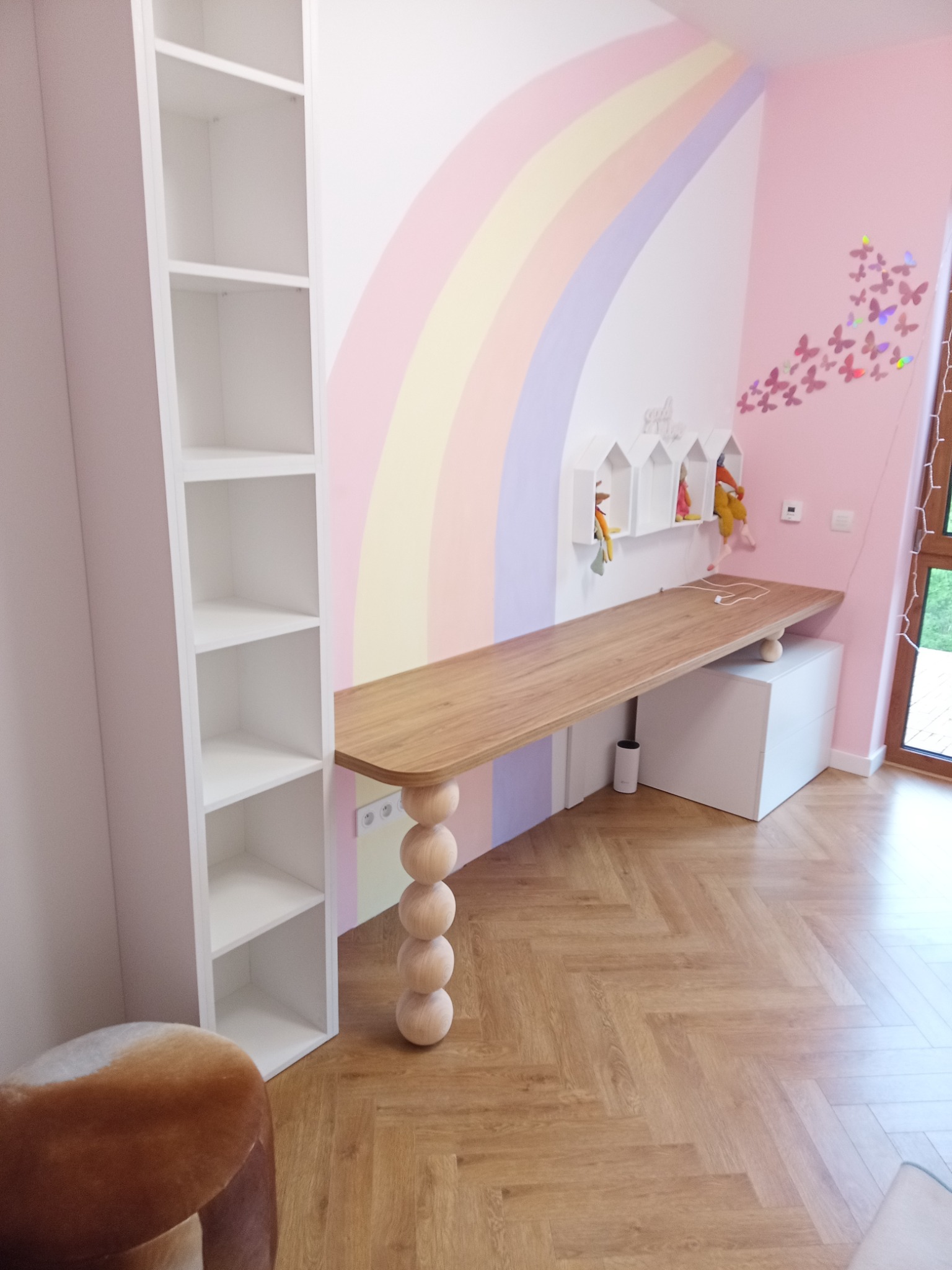 Pokój dziecięcy z biurkiem z blatem drewnianym na toczonych nogach, regałem i tęczową ścianą. Wnętrze w pastelowych kolorach.