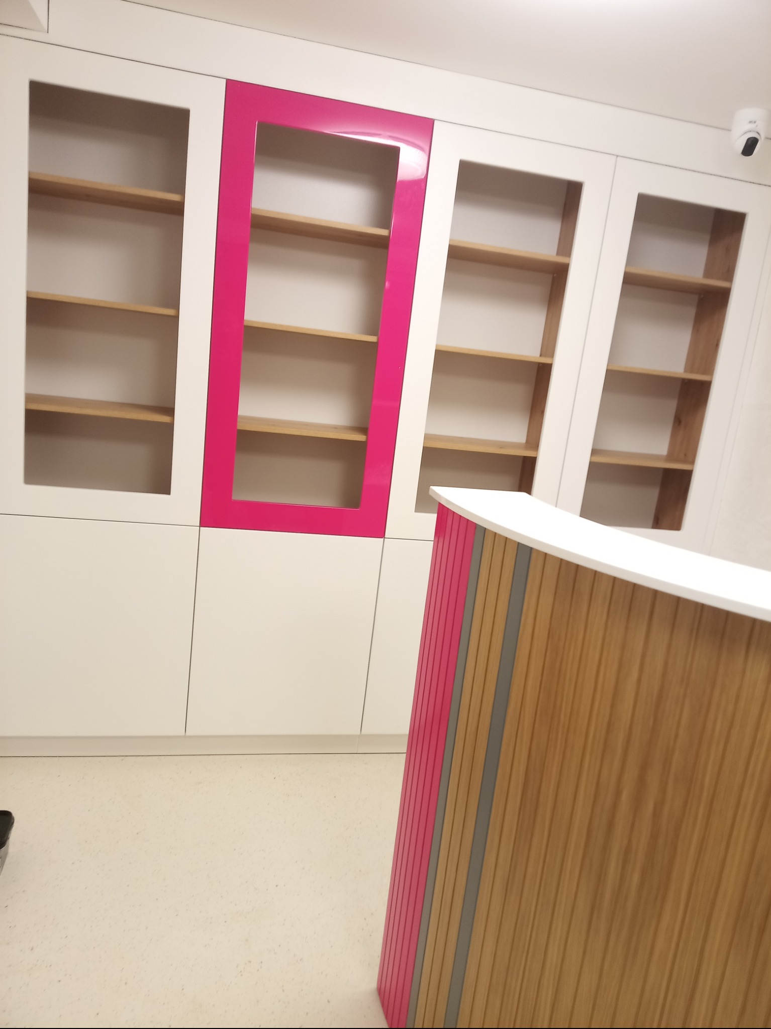 Białe szafy z przeszklonymi drzwiami i drewnianymi półkami, akcentowane różową ramą, oraz drewniana lada recepcyjna z różowymi elementami.