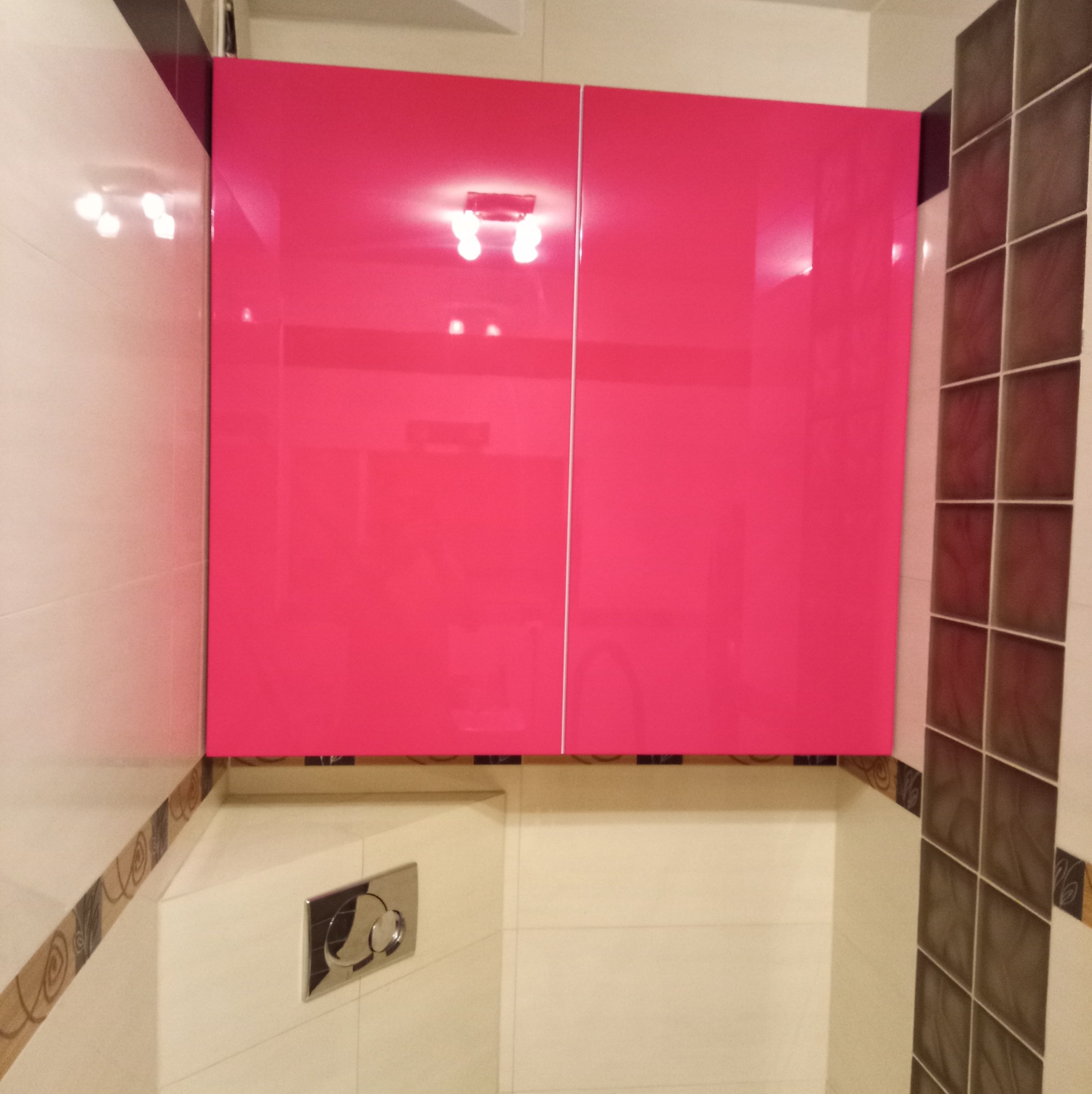 Różowa szafka łazienkowa z połyskiem, zamontowana nad toaletą. Widoczna armatura, białe i brązowe płytki. Nowoczesny design, minimalizm i funkcjonalność.