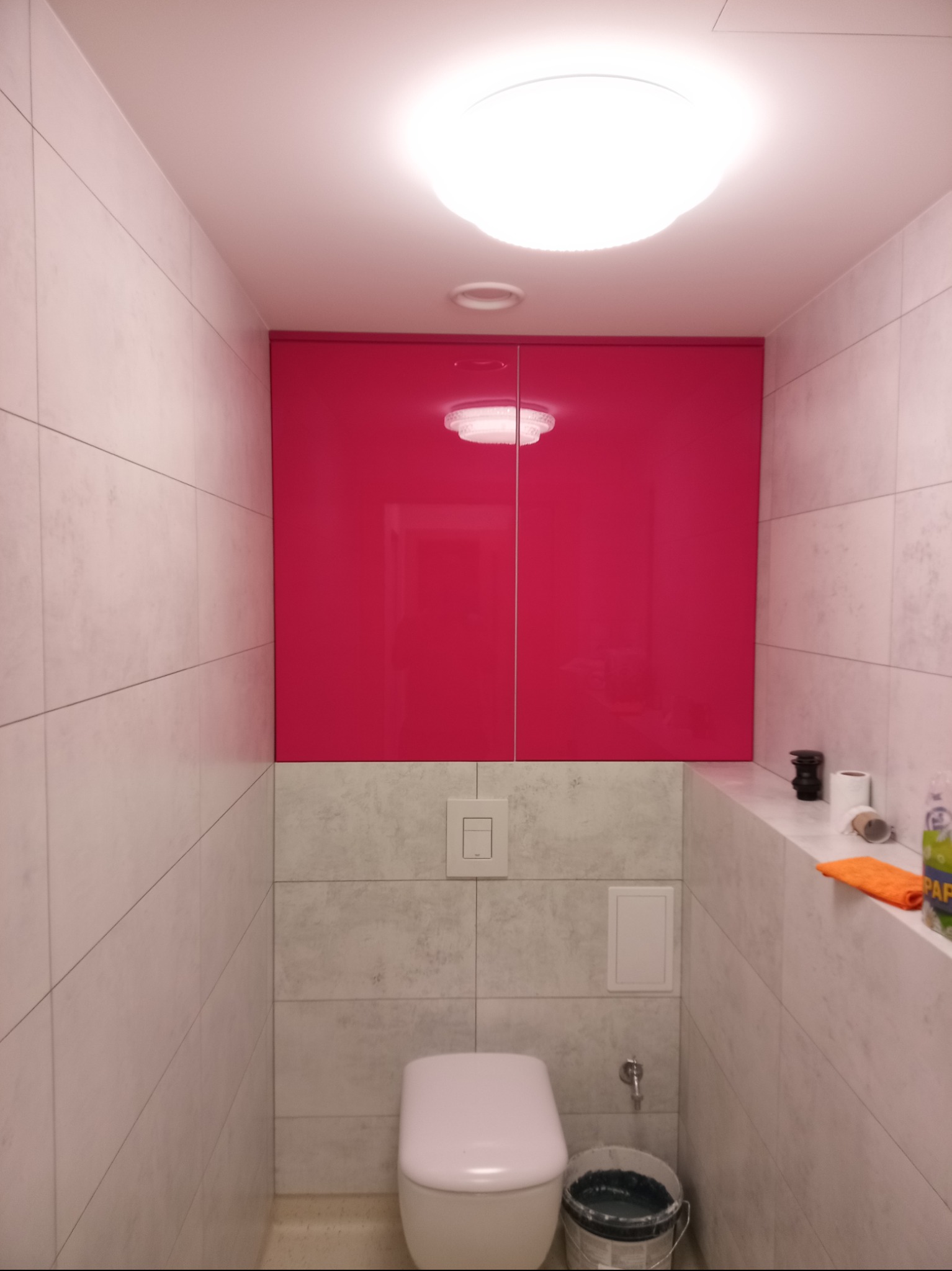 Nowoczesna toaleta z szafką w intensywnym różu nad zabudowaną spłuczką. Białe płytki imitujące kamień, okrągła lampa sufitowa. Widoczny fragment półki i akcesoria.
