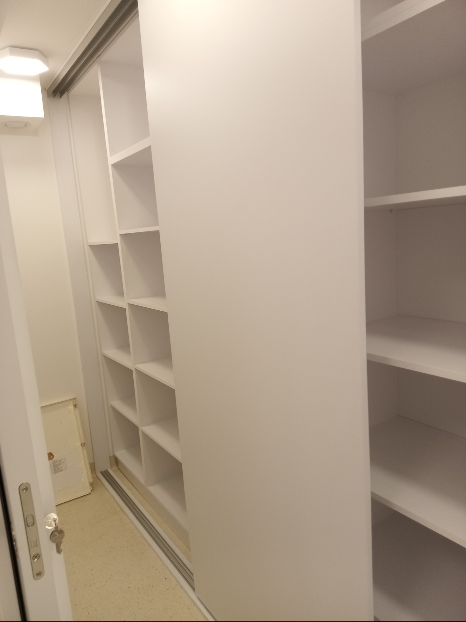 Biała szafa wnękowa z przesuwnymi drzwiami, widoczne półki. Ujęcie z perspektywy otwartych drzwi wejściowych do pomieszczenia.