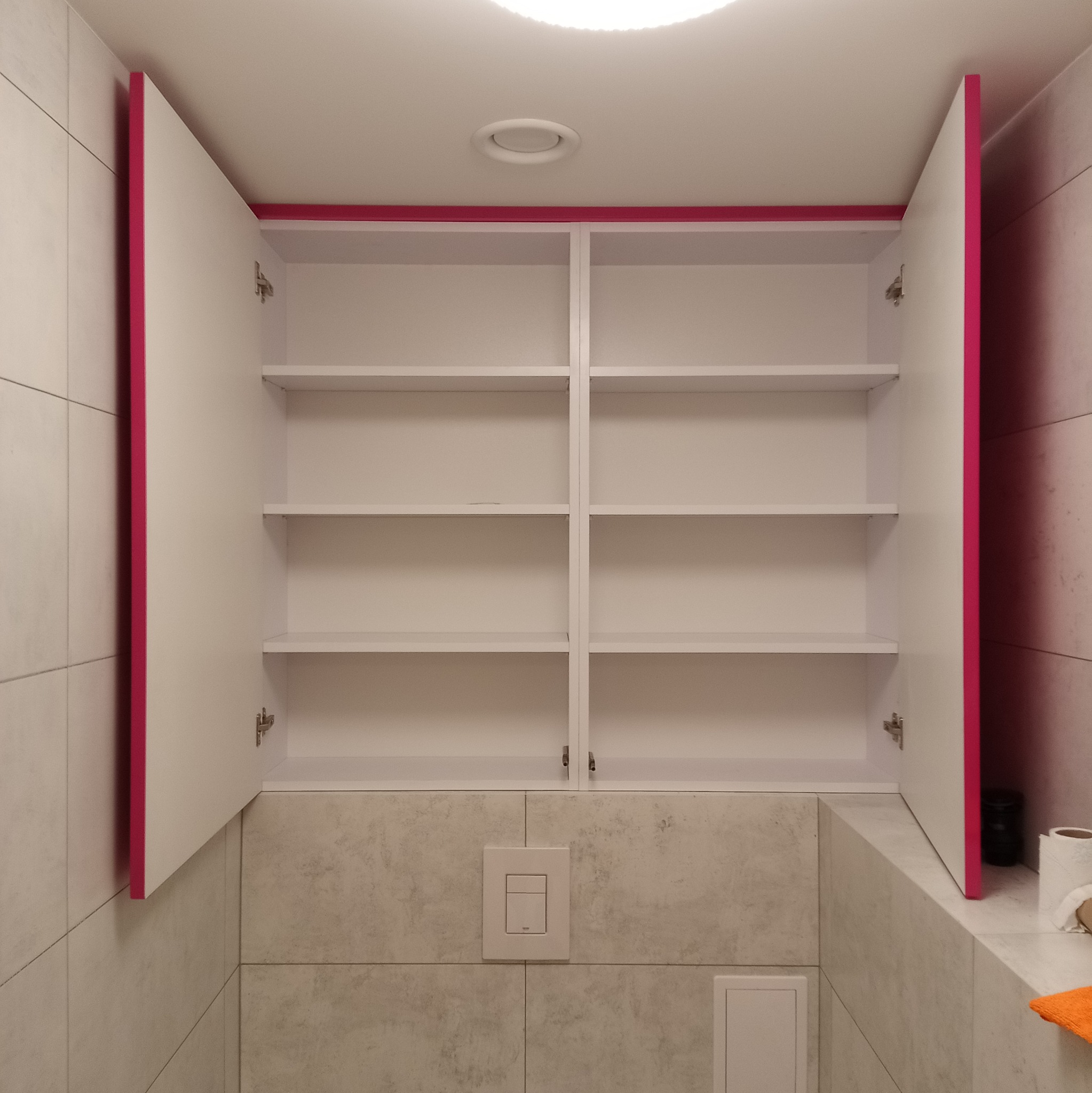 Otwarta szafka w łazience z białymi półkami i różowymi obramowaniami drzwi, zamontowana nad toaletą. Widoczne płytki na ścianach i włącznik spłuczki.