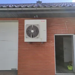 Biała jednostka zewnętrzna klimatyzacji Toshiba zamontowana na ceglanej ścianie budynku, widoczna brama garażowa Wiśniowski po lewej i otwarte drzwi do pomieszczenia po prawej.