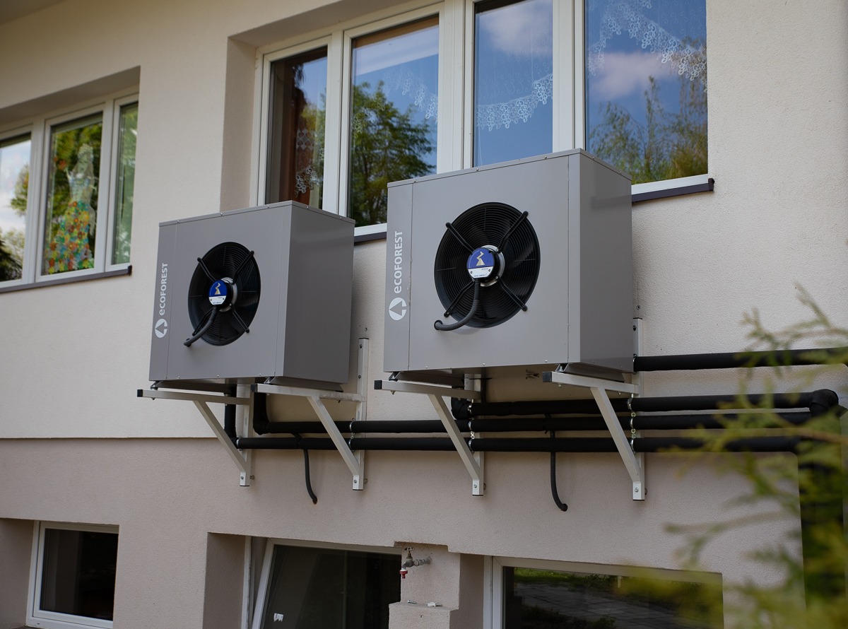 Dwa szare moduły zewnętrzne pompy ciepła Ecoforest zamontowane na wspornikach na elewacji budynku, widoczne czarne rury instalacji i okna z odbiciem nieba.
