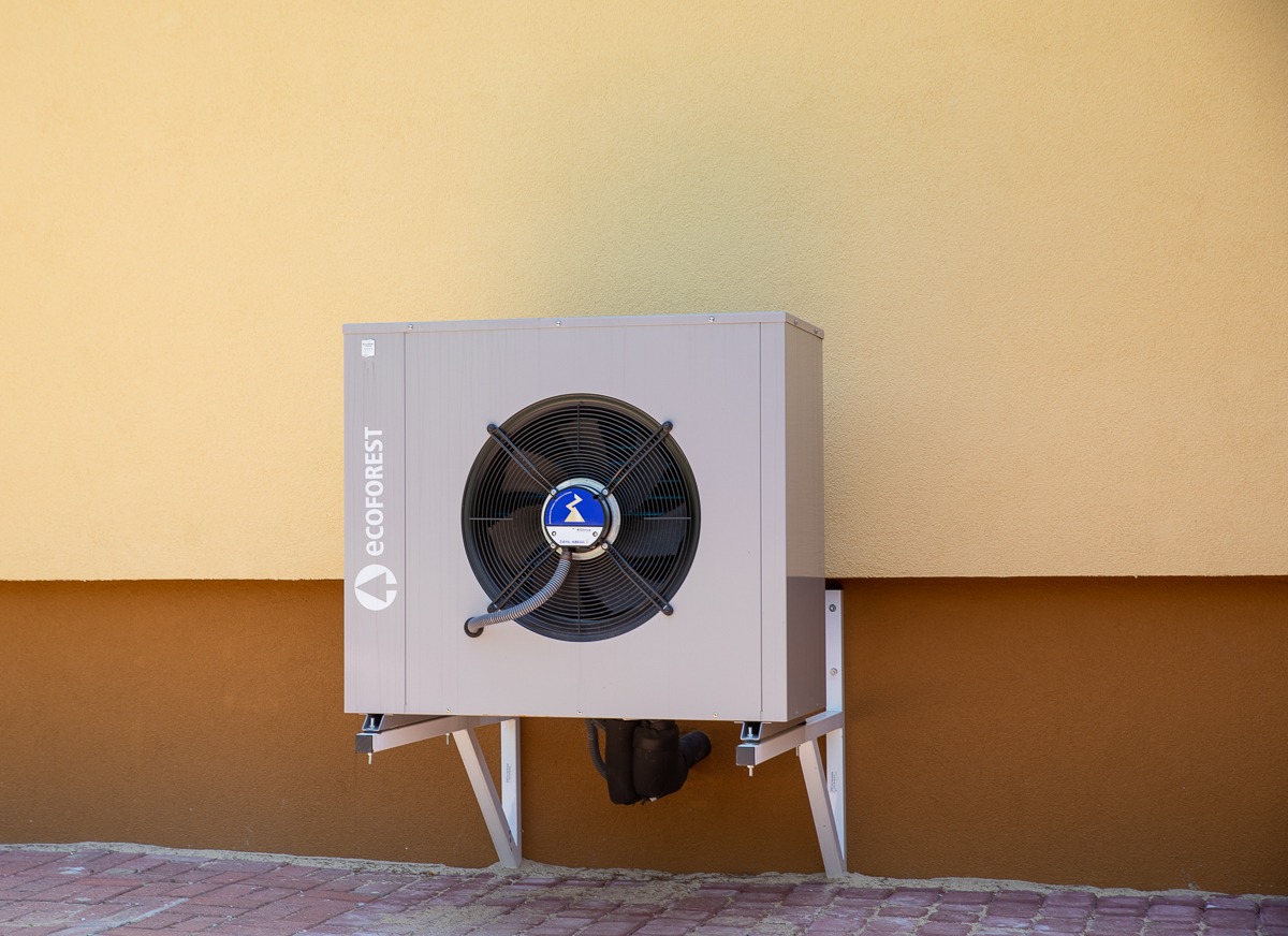 Zewnętrzna jednostka pompy ciepła marki Ecoforest, zamontowana na wspornikach przy elewacji budynku z dwukolorową fasadą, widok z przodu.