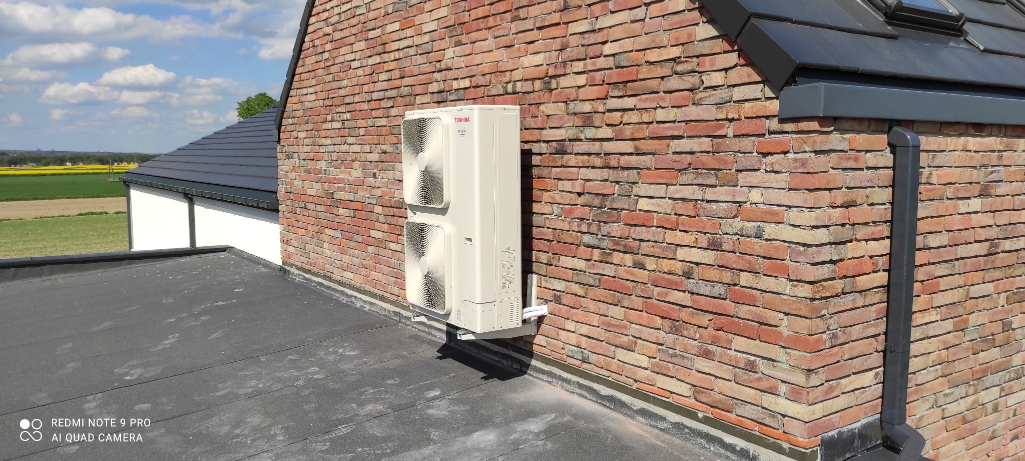 Biała jednostka zewnętrzna klimatyzacji Toshiba zamontowana na ceglanej ścianie budynku z widocznym dachem pokrytym papą i fragmentem pola w tle.