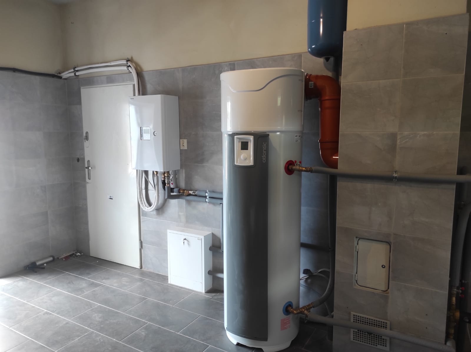 Nowoczesna instalacja grzewcza z zasobnikiem ciepłej wody Atlantic, widoczna w pomieszczeniu z szarymi płytkami i białymi rurami oraz dodatkowym piecykiem na ścianie.