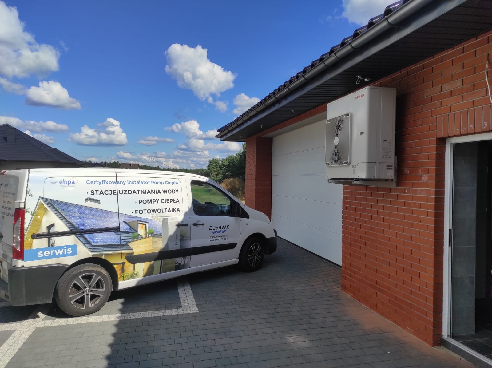 Zewnętrzna jednostka pompy ciepła Toshiba zamontowana na ceglanej ścianie budynku, obok białej bramy garażowej; na pierwszym planie samochód firmowy z reklamą usług instalacyjnych, w tle błękitne...