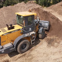 PHUiP INTERCO - Żółta ładowarka kołowa XCMG nabiera materiał z hałdy, widoczne ślady opon na piaszczystym podłożu, drzewa w tle.