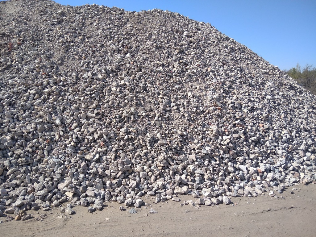 Duża pryzma szarego kruszywa z domieszką fragmentów cegieł pod błękitnym niebem, widoczna krawędź terenu.