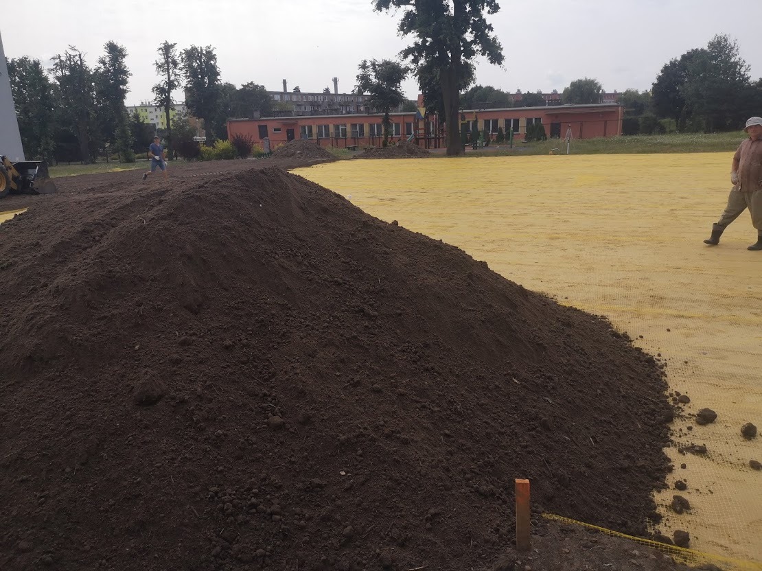 Kopuła ciemnej, wilgotnej ziemi na żółtej macie geotekstylnej, w tle pracownik i budynek szkolny, widoczne pomarańczowe paliki wyznaczające teren.
