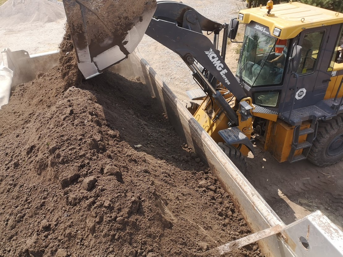 Żółta ładowarka XCMG Europe nasypuje ziemię ogrodową do metalowej skrzyni na przyczepie.