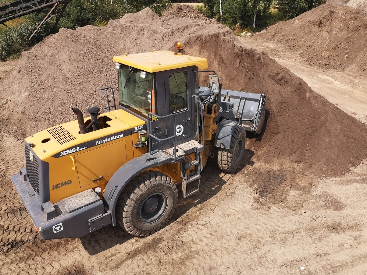 Żółta ładowarka kołowa XCMG nabiera materiał z hałdy, widoczne ślady opon na piaszczystym podłożu, drzewa w tle.