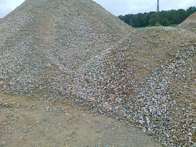 Stos szarego kruszywa o różnej granulacji, z widocznym śladem opon na szczycie, na tle zieleni i słupa energetycznego.