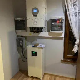 Domowa instalacja fotowoltaiczna: biały inwerter z wyświetlaczem, biały magazyn energii na kółkach z zieloną diodą, rozdzielnice AC i DC na ścianie w rogu pokoju z drewnianą podłogą i oknem...