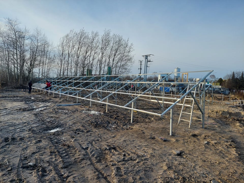 Montaż metalowej konstrukcji wsporczej pod panele słoneczne na błotnistym terenie, widoczni dwaj pracownicy w oddali, jeden w czerwonej kurtce, drabina oparta o konstrukcję, w tle budynki i słupy...