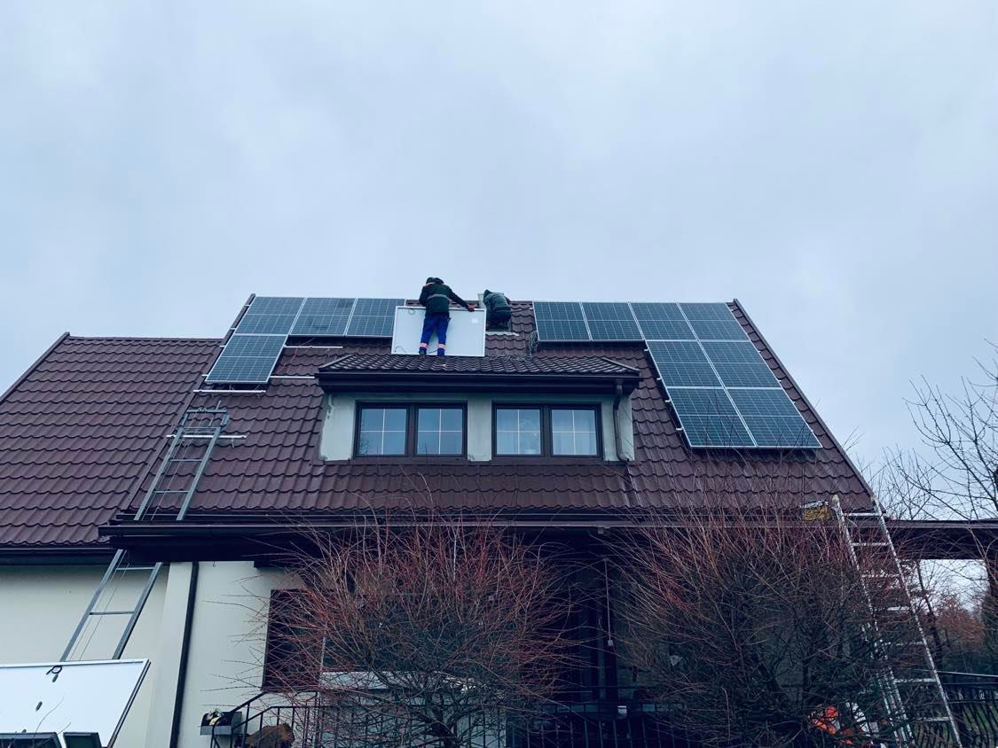 Montaż paneli słonecznych na dachu domu z brązową dachówką; dwóch instalatorów w odzieży roboczej umieszcza panel na wysokości, drabiny opierają się o ścianę i dach.