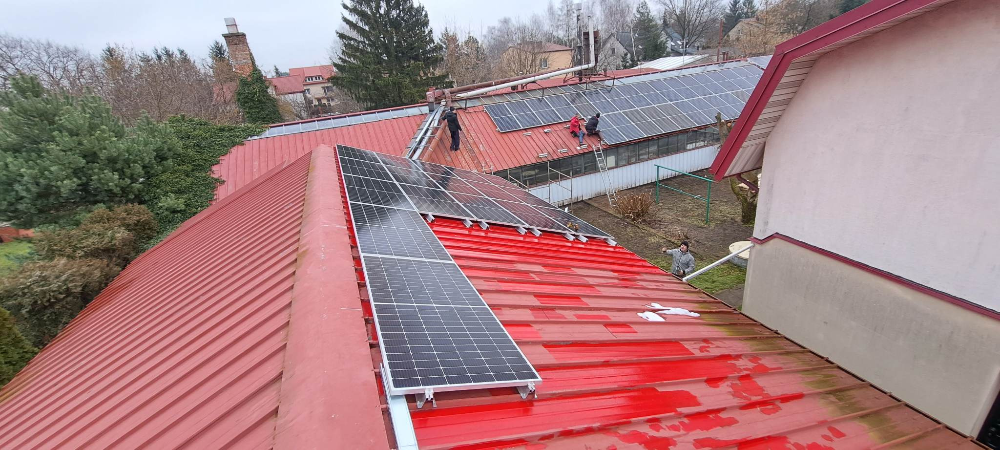 Montaż paneli słonecznych na czerwonym, blaszanym dachu budynku; widoczni pracownicy na drabinie i dachu, instalujący panele w pochmurny dzień.