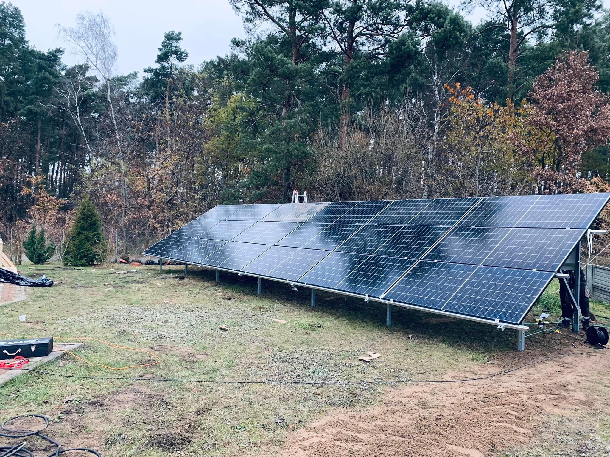 Panel słoneczny na metalowej konstrukcji, montowany na trawiastym terenie z widocznymi śladami prac ziemnych, w tle las mieszany pod pochmurnym niebem.