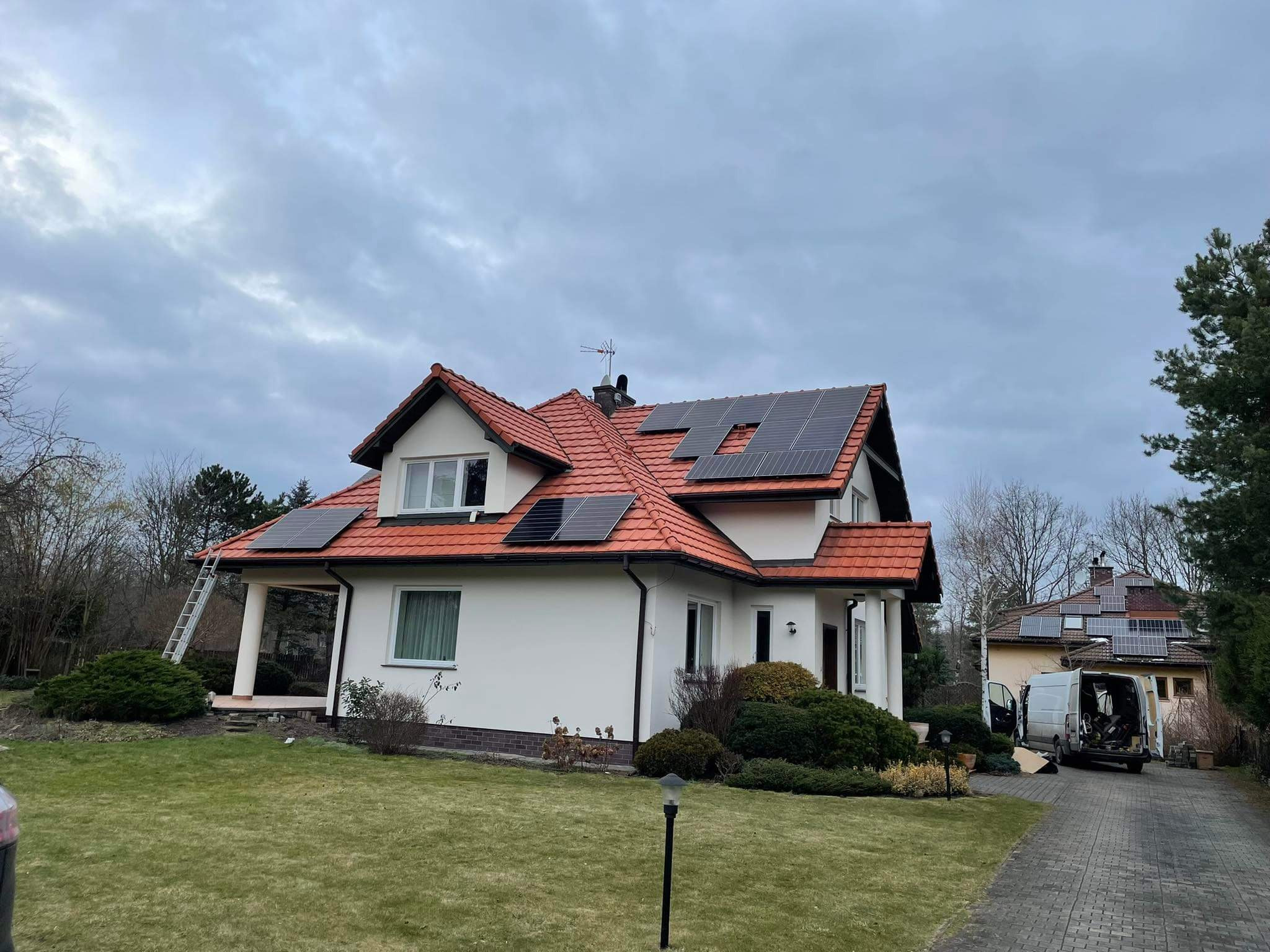Dom jednorodzinny z czerwonym dachem, na którym zamontowano czarne panele fotowoltaiczne; drabina oparta o ścianę, a w tle zaparkowany biały van z otwartymi drzwiami.