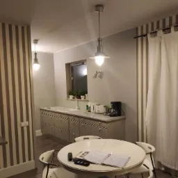 Ustawna kuchnia z szarymi szafkami, okrągłym stołem z białymi krzesłami i wiszącymi lampami w stylu industrialnym, na blacie ekspres do kawy i toster.