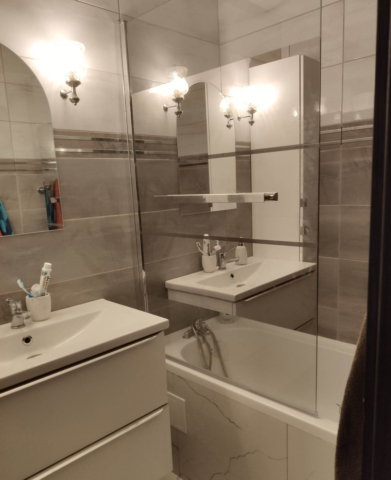 Umywalki z szafkami, lustro i wanna za szklaną ścianką w łazience z szarymi płytkami, oświetlone kinkietami z kloszami.