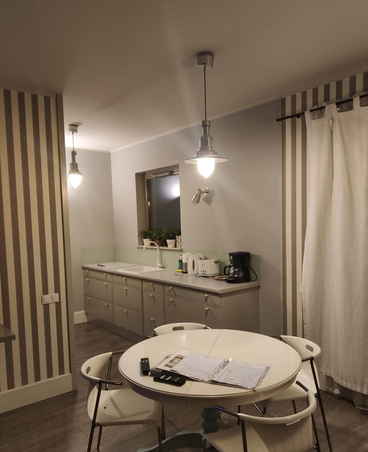 Ustawna kuchnia z szarymi szafkami, okrągłym stołem z białymi krzesłami i wiszącymi lampami w stylu industrialnym, na blacie ekspres do kawy i toster.