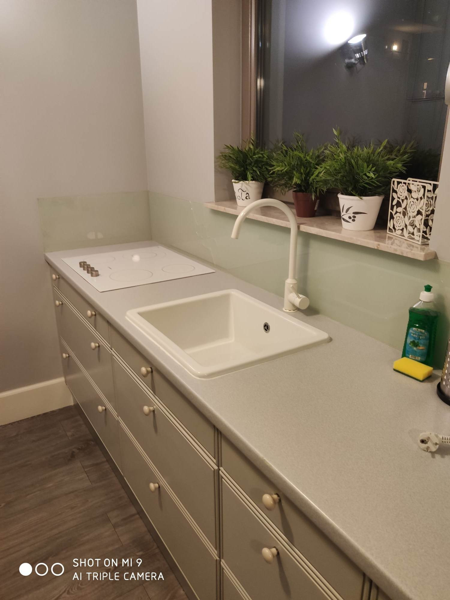 Jasna kuchnia z białym zlewem i płytą indukcyjną, okno z roślinami w tle, minimalistyczny design.