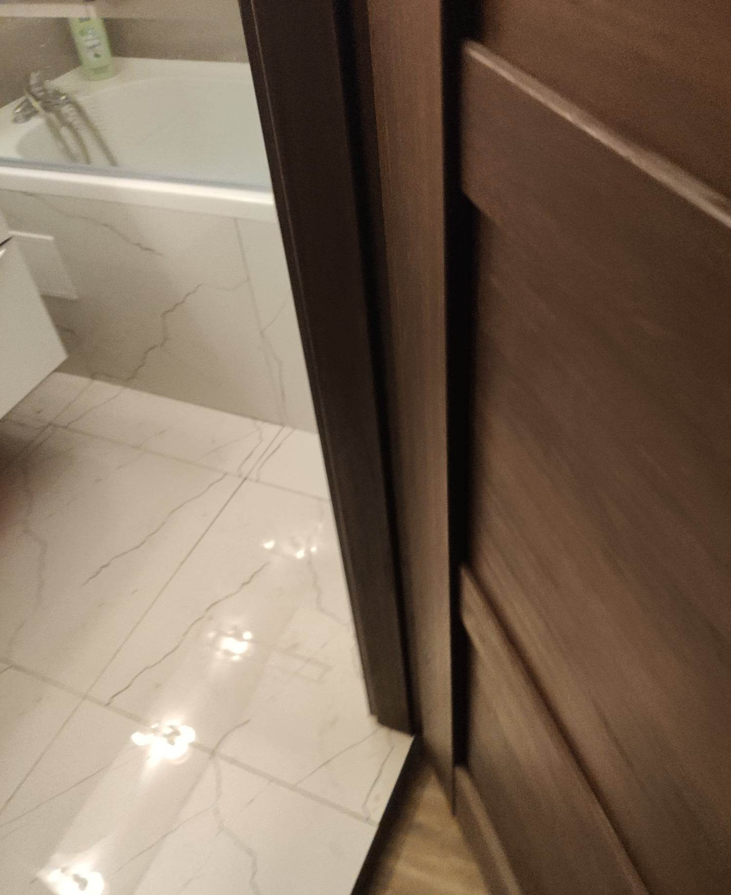 Fragment łazienki z widoczną białą wanną, marmurową posadzką i częściowo otwartymi, brązowymi drzwiami.