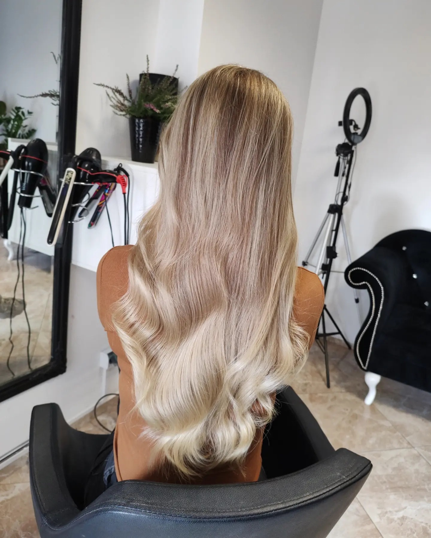 Blond włosy z delikatnymi falami, widok z tyłu, na fotelu w salonie, z widocznym oświetleniem pierścieniowym i akcesoriami do stylizacji włosów.