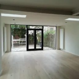 Wnętrze pokoju po remoncie z jasną drewnianą podłogą, białymi ścianami i dużymi oknami z czarnymi ramami wychodzącymi na ogród z kwitnącymi drzewami. Widoczne grzejniki na ścianach i rusztowanie za...