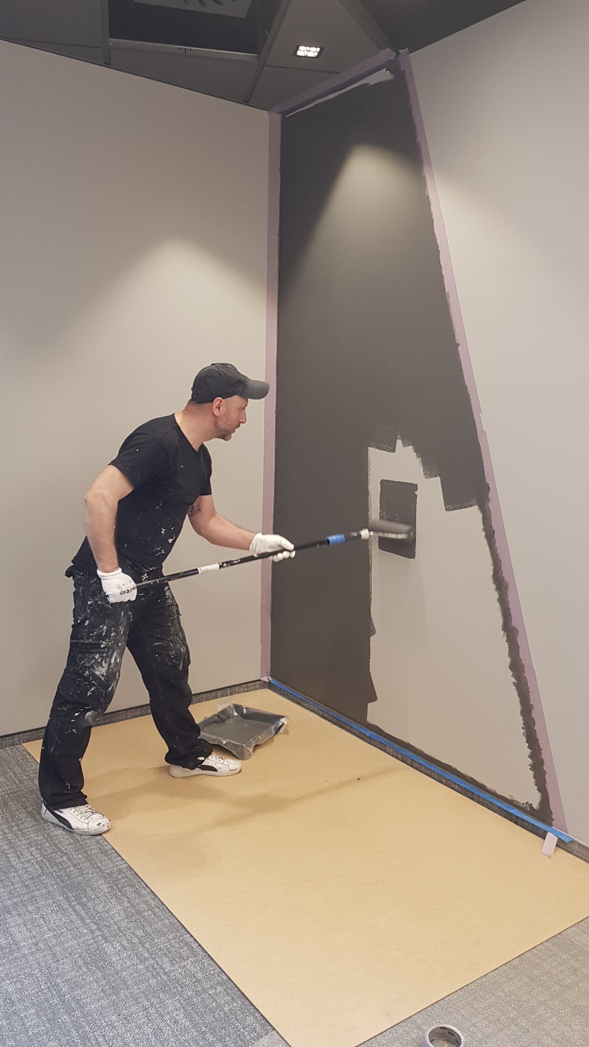 Mężczyzna w czarnym ubraniu i białych rękawicach maluje szarą farbą ścianę w biurze za pomocą wałka na długim kiju, podłoga zabezpieczona kartonem i taśmą malarską, widoczne ślady farby na ubraniu.