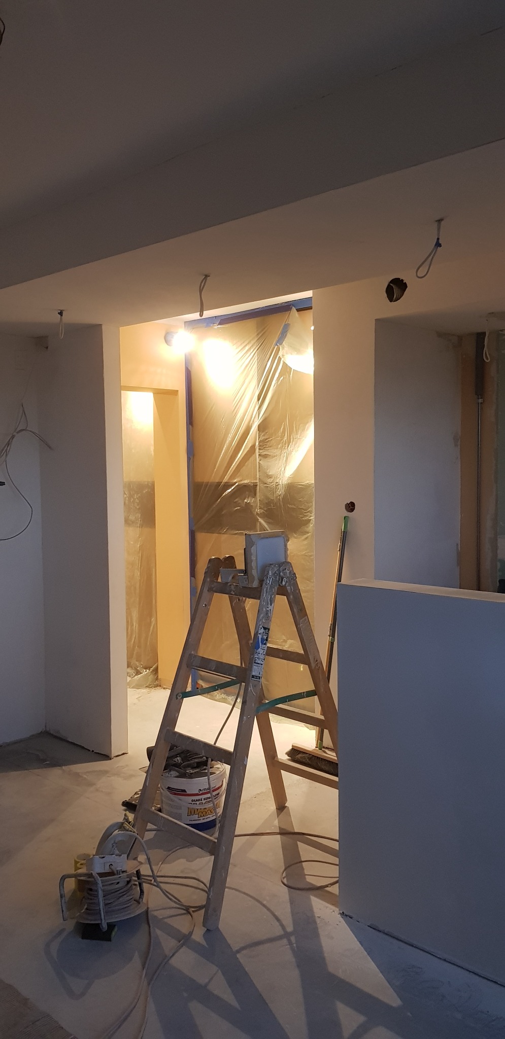 Przygotowania do prac wykończeniowych: drewniana drabina z wiadrem z masą szpachlową i narzędziami, pomieszczenie zabezpieczone folią malarską, widoczny przedłużacz elektryczny.