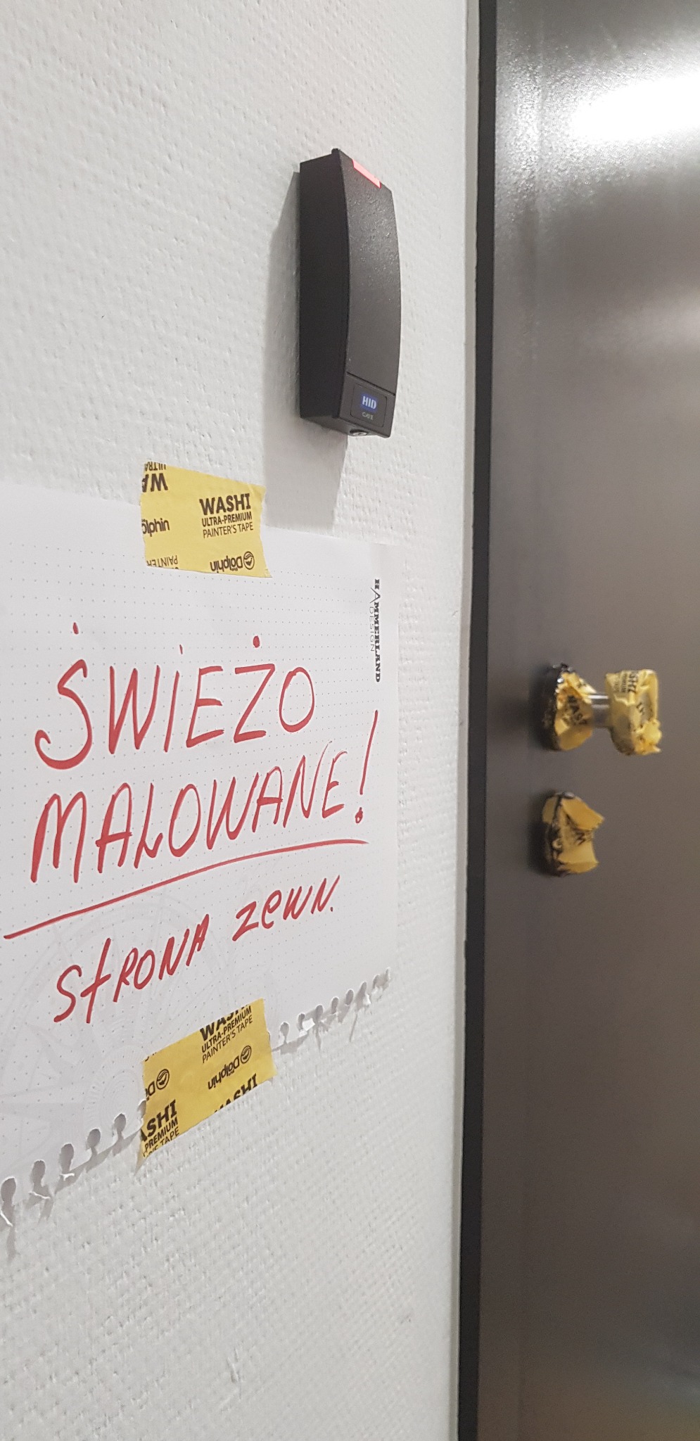 Świeżo pomalowana ściana z kartką 'Świeżo malowane! strona zewn.' przymocowaną taśmą malarską, obok czytnik kart dostępu i fragment drzwi zabezpieczonych taśmą malarską.