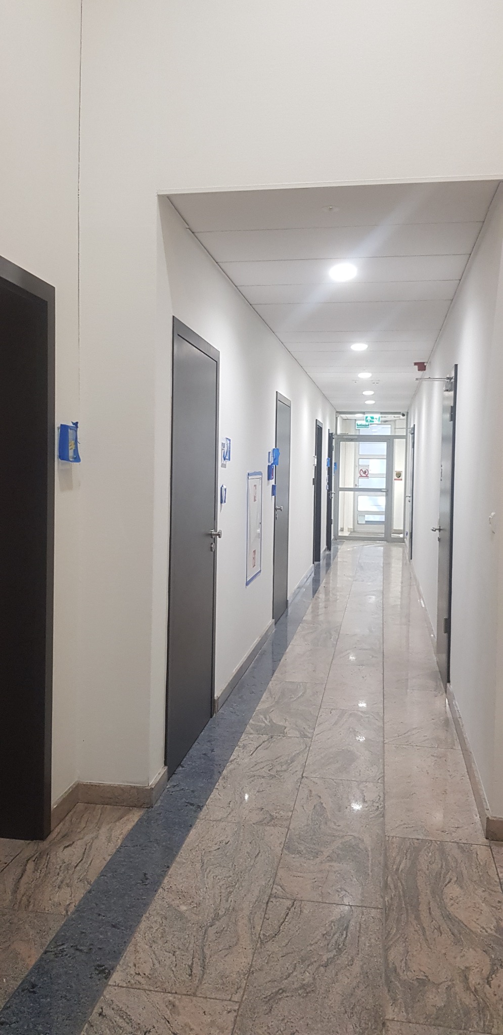 Długi korytarz biurowy z białymi ścianami, szarymi drzwiami i marmurową podłogą z ciemniejszym pasem po środku.