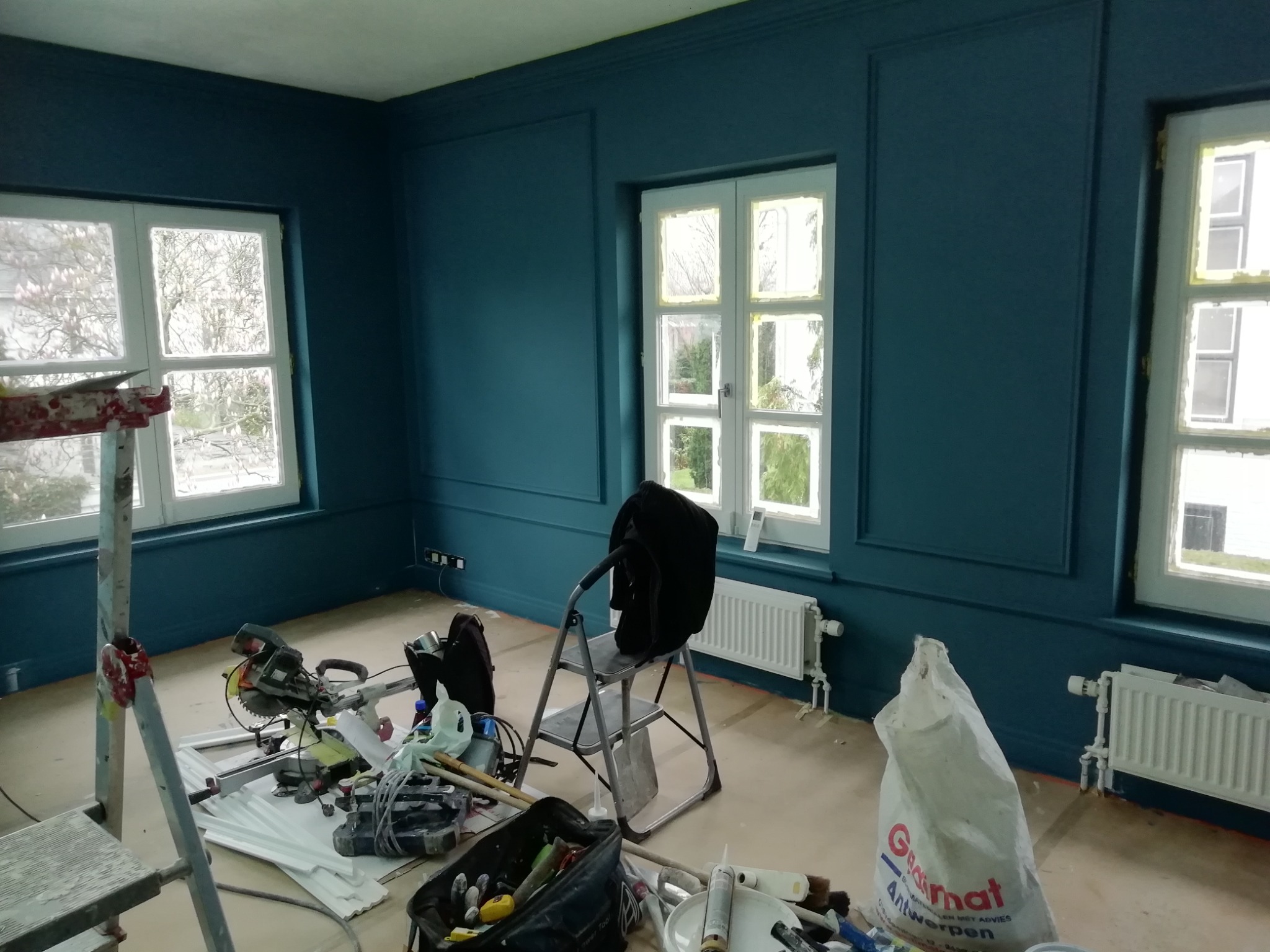 Pokój w trakcie remontu, ściany pomalowane na ciemny, morski kolor, białe okna zabezpieczone taśmą malarską, widoczne narzędzia malarskie, drabina i worki z materiałami budowlanymi.