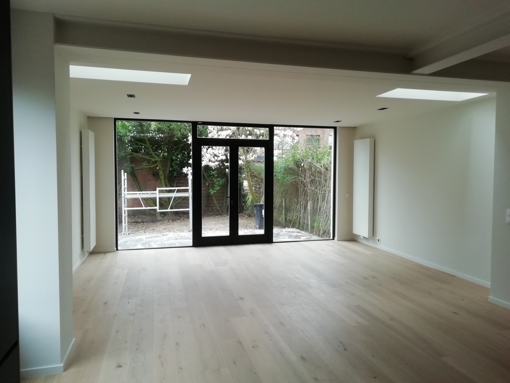 Wnętrze pokoju po remoncie z jasną drewnianą podłogą, białymi ścianami i dużymi oknami z czarnymi ramami wychodzącymi na ogród z kwitnącymi drzewami. Widoczne grzejniki na ścianach i rusztowanie za...