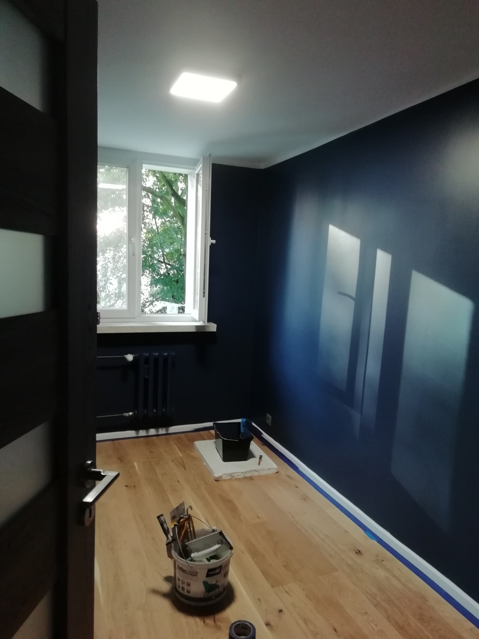 Pomieszczenie po malowaniu na ciemny granat, widoczne narzędzia malarskie, zabezpieczona listwa przypodłogowa taśmą malarską, drewniana podłoga, okno z widokiem na zieleń.
