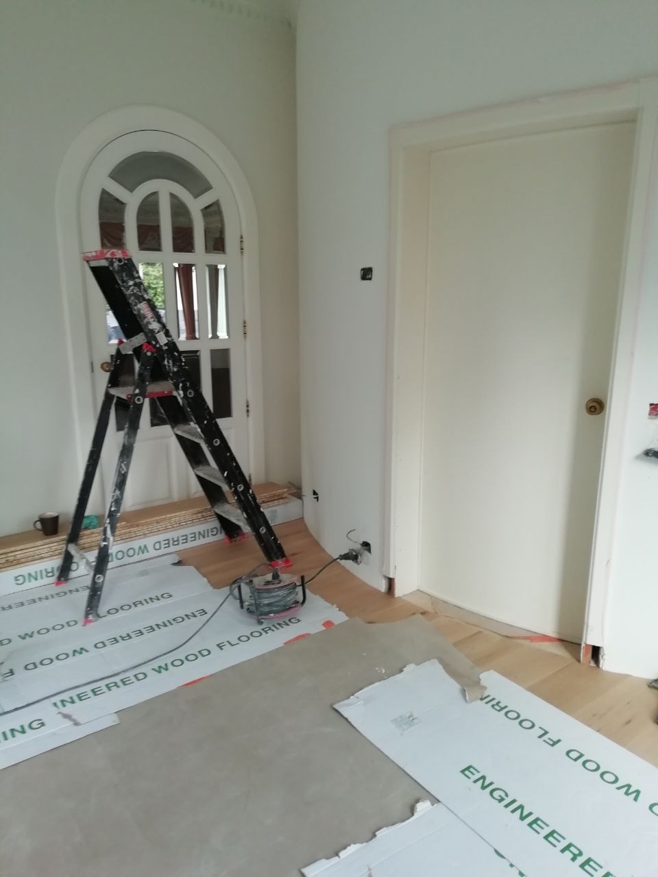 Pokój w trakcie remontu: świeżo pomalowane białe ściany, rozłożone panele podłogowe z widocznym napisem 'Engineered Wood Flooring', drabina przy łukowatych drzwiach, widoczny kabel zasilający.