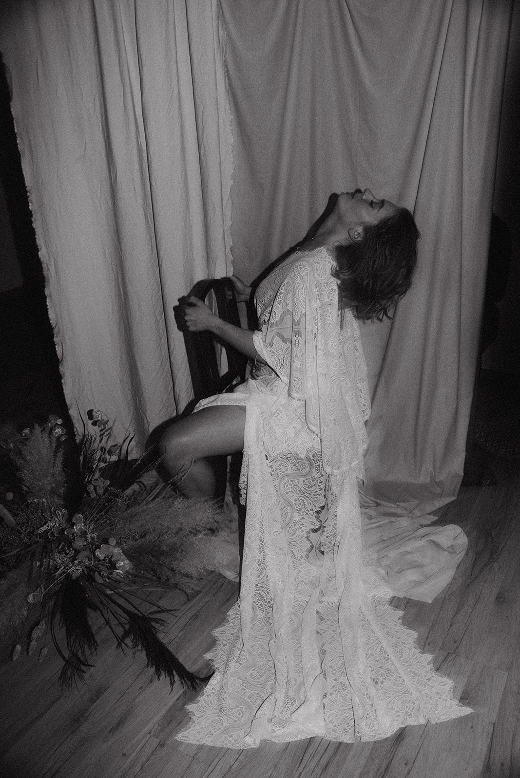 Kobieta w koronkowej narzutce, siedząca na krześle z głową odchyloną do tyłu, obok bukiet suchych traw i kwiatów, czarno-biała fotografia.