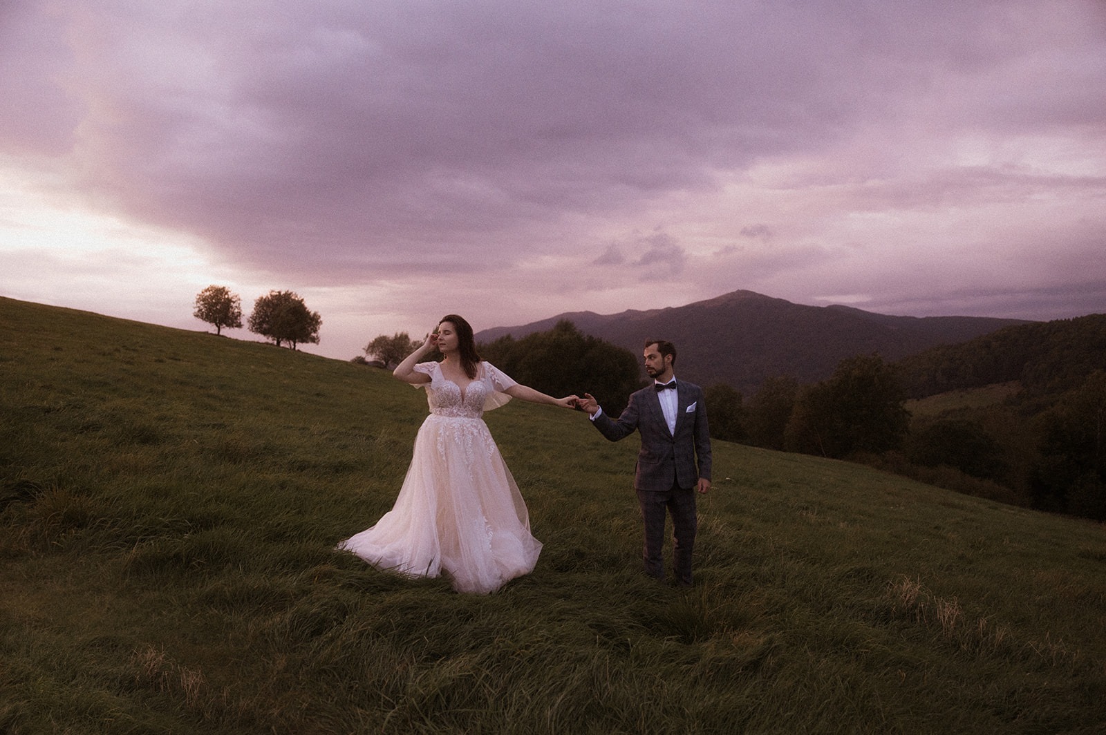 Para młoda trzymająca się za ręce na łące o zmierzchu, góry w tle.