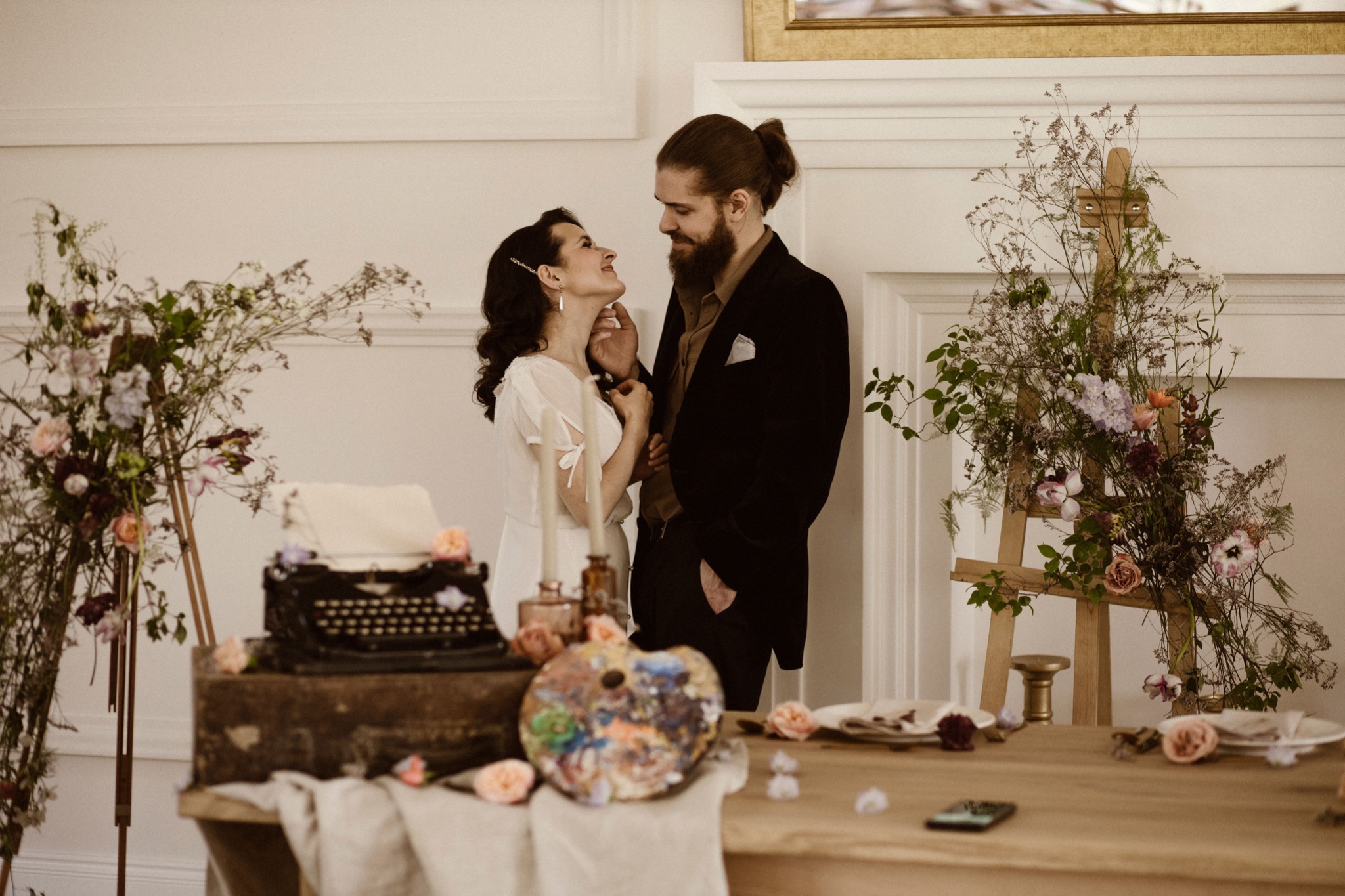 Para zakochanych w stylizowanej scenerii sesji ciążowej: kobieta w zwiewnej sukience i mężczyzna w marynarce, otoczeni kwiatami, starą maszyną do pisania i paletą malarską na drewnianym stole.