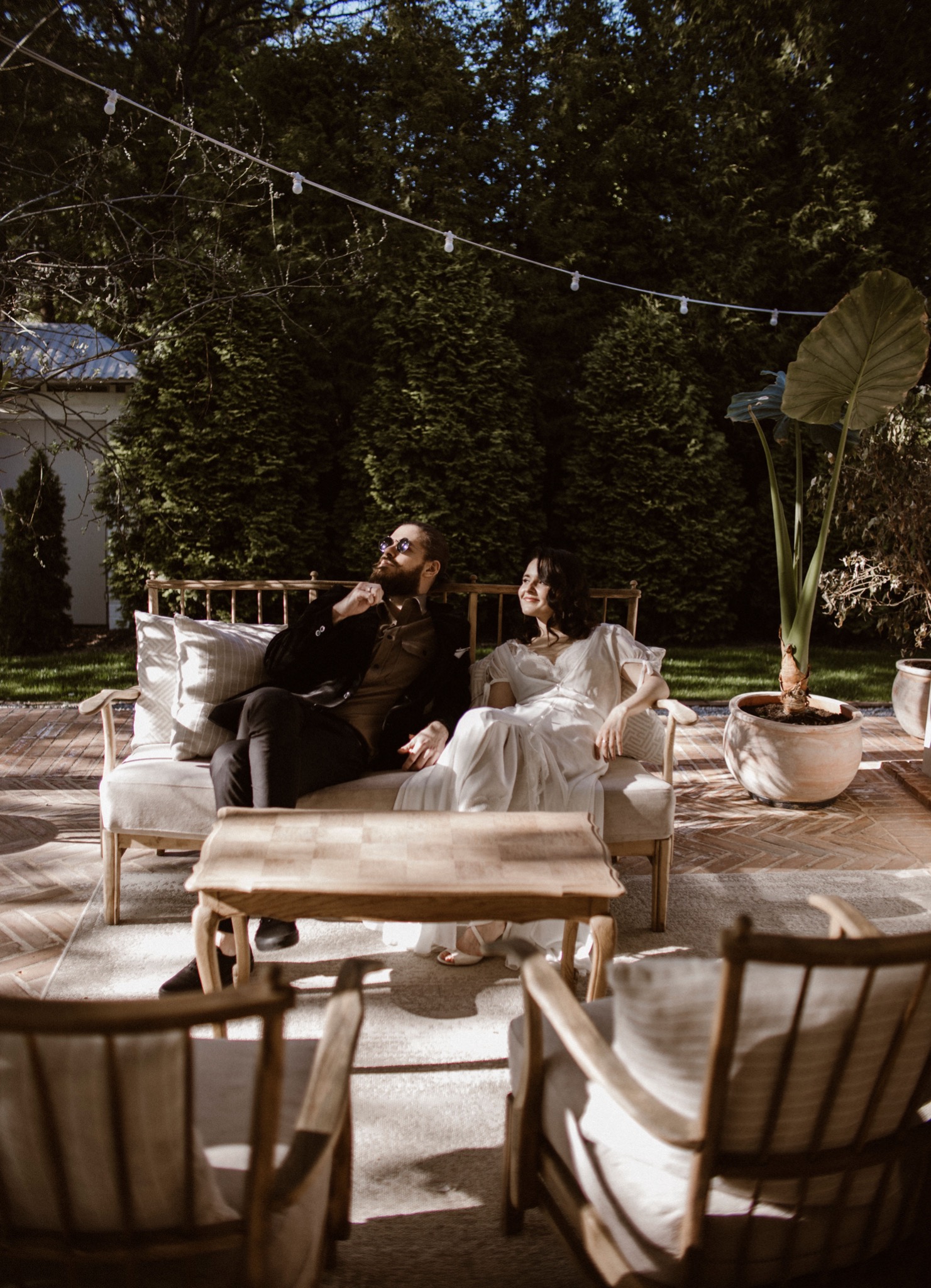 Para w eleganckich strojach relaksuje się na tarasie z drewnianymi meblami w otoczeniu zieleni i dekoracyjnych światełek.