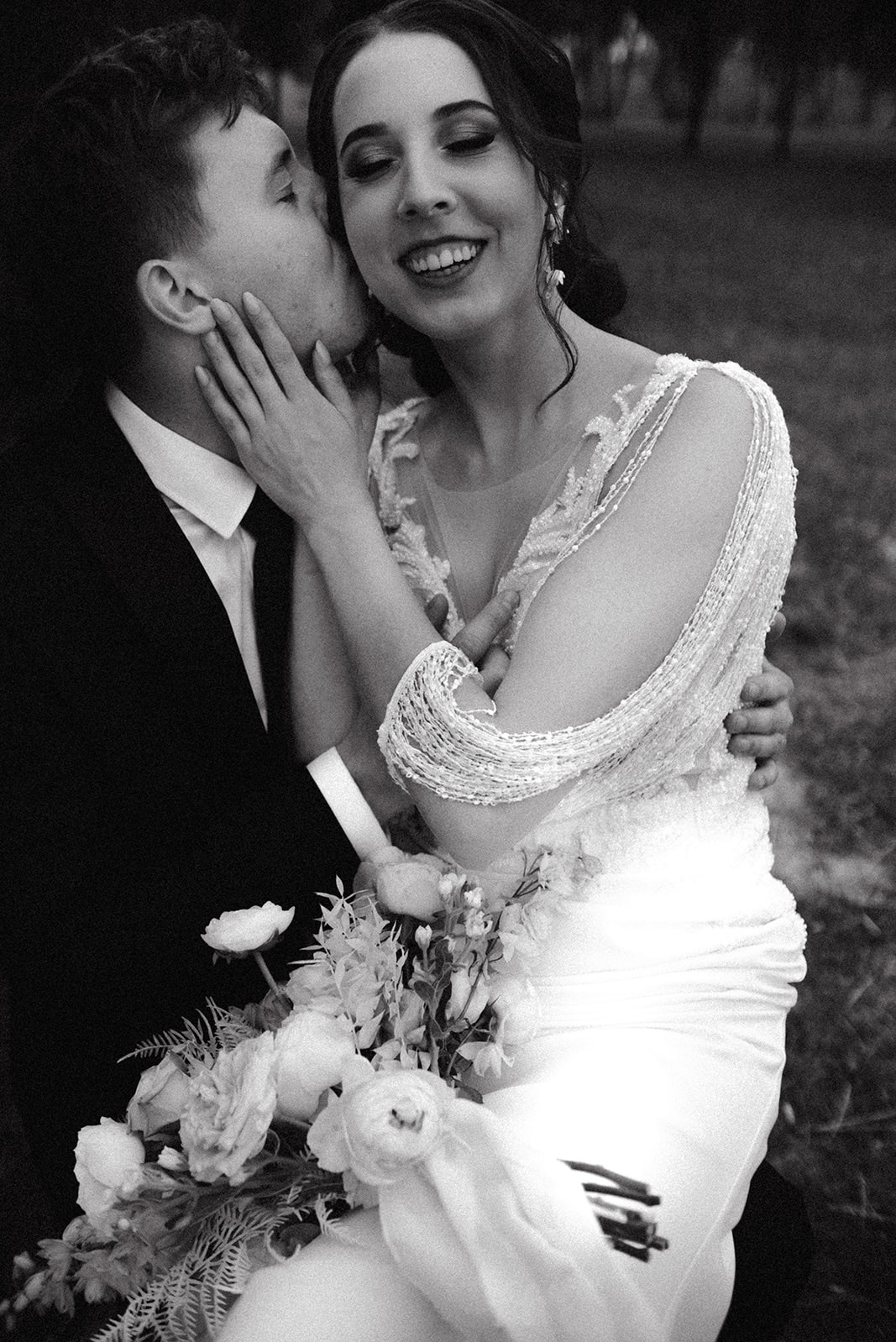 Czarno-białe zdjęcie uśmiechniętej kobiety w sukni ślubnej, obejmowanej przez mężczyznę w garniturze, który całuje ją w policzek; bukiet białych kwiatów w dolnej części kadru.