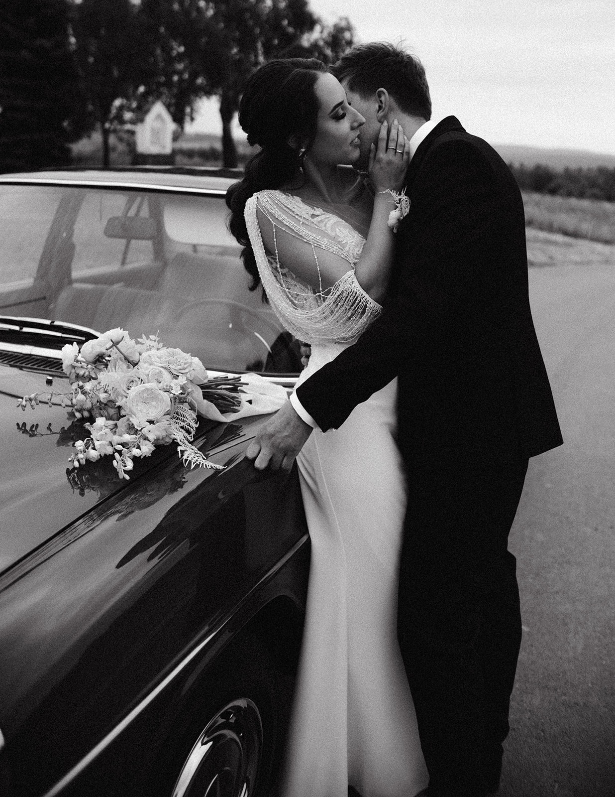 Czarno-białe zdjęcie nowożeńców przy klasycznym samochodzie: panna młoda w sukni z frędzlami i bukietem na masce, pan młody w czarnym garniturze.