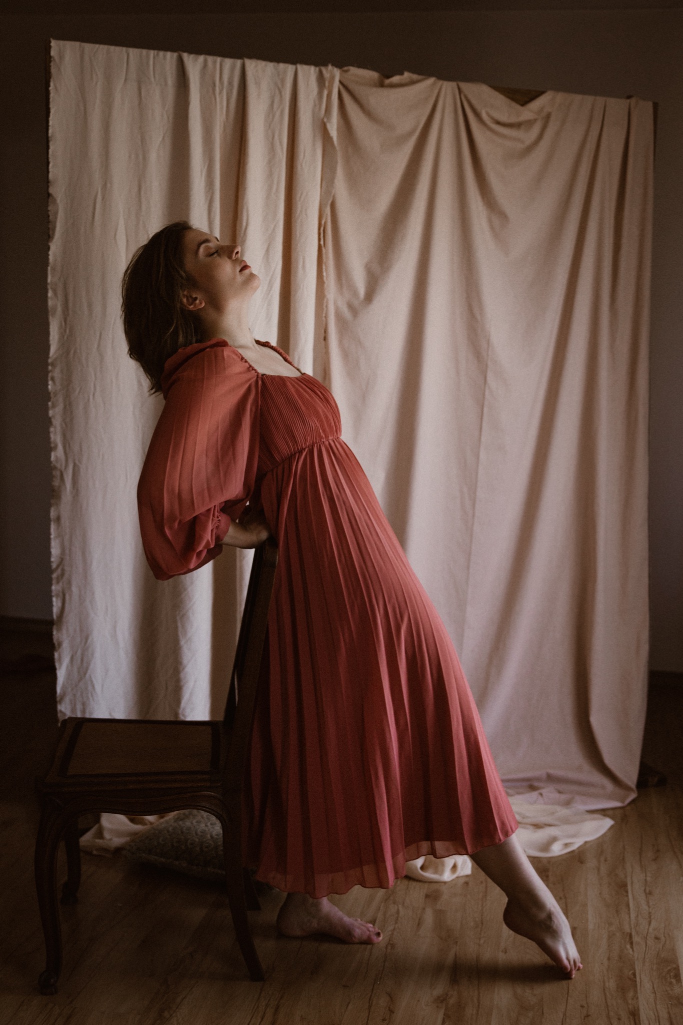 Kobieta w plisowanej sukience w kolorze terakoty, oparta plecami o krzesło, z głową odchyloną do tyłu, pozuje na tle beżowej tkaniny